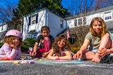 Three little girls and a boy sitting on the driveway outside a suburban home drawing with chalk