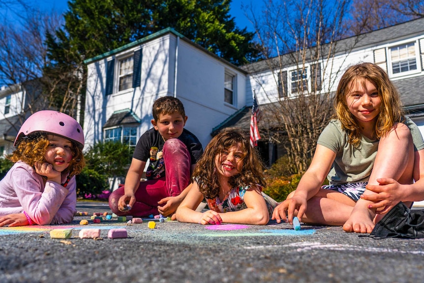 Three little girls and a boy sitting on the driveway outside a suburban home drawing with chalk