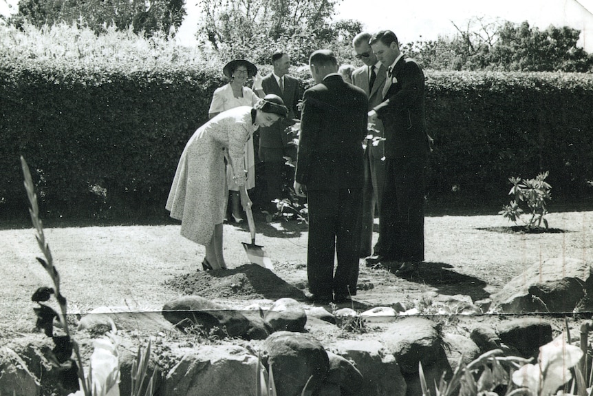 Queen Elizabeth II digs a hole with a shovel while others stand near her in a garden.