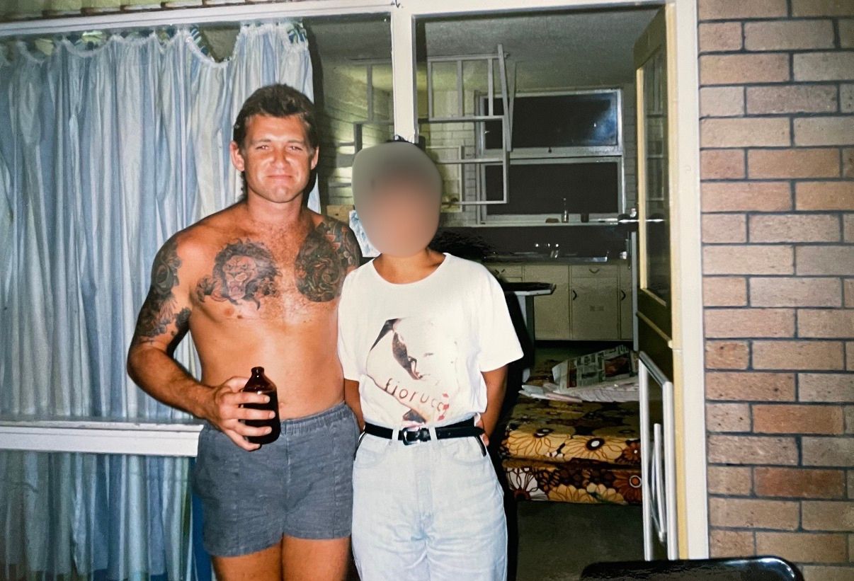 A shirtless man with tattoos holding a beer and standing with a woman whose face is blurred.