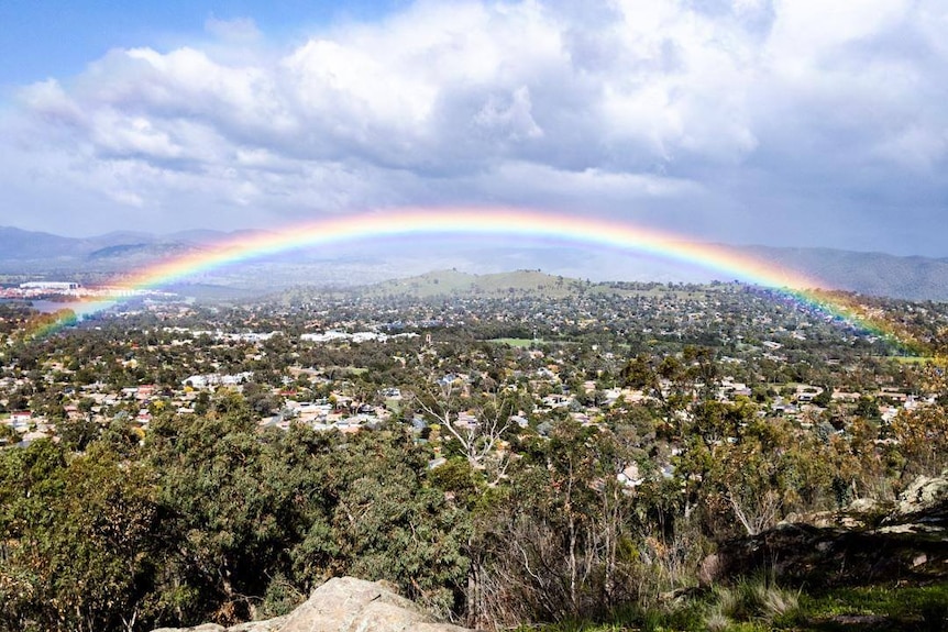 A rainbow over Canberra's hills with houses in the background.