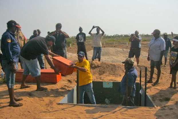 A group of Indigenous people in a sandy area, with some standing in a square hole, about to put some wooden boxes in it.