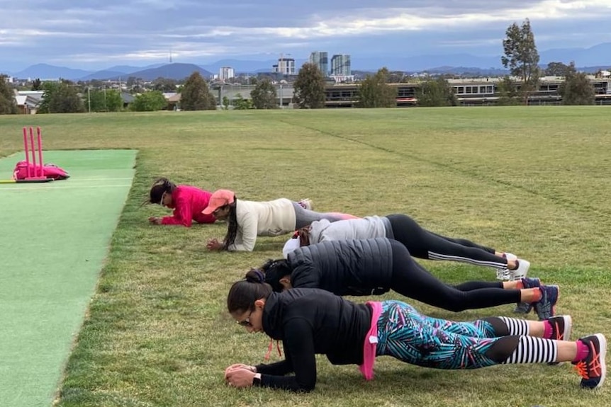 A row of five women plank on a cricket field.