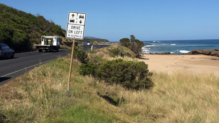cars and truck on a road beside the ocean with a road sign in the foreground saying drive on left