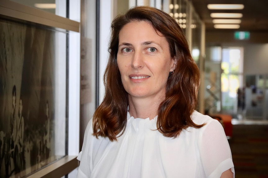 A woman with brown hair and wearing a white blouse looking serious, standing in a hallway. 