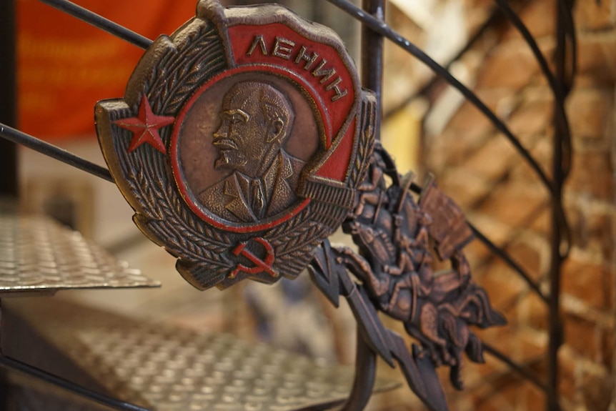 A metal shield shows the bust of Lenin looking to the left encircled by a wreath of wheat with a start and hammer and sickle