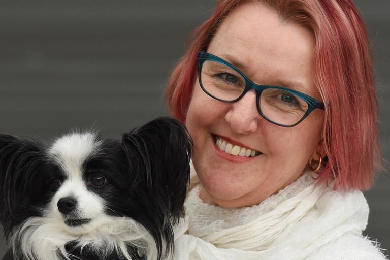 A lady with light red hair and glasses holds a small black and white dog.