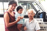 A young woman holding a toddler, stands with an elderly woman. Both women are smiling.