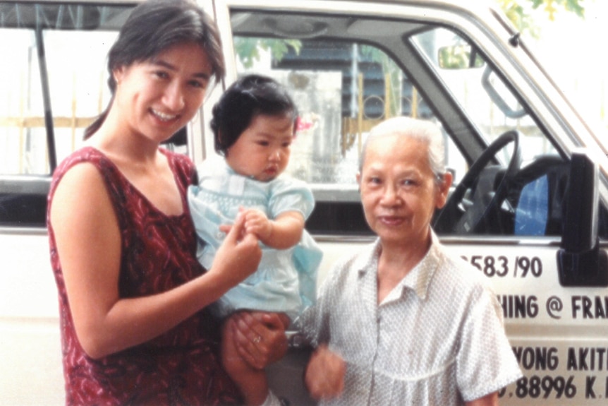 A young woman holding a toddler, stands with an elderly woman. Both women are smiling.