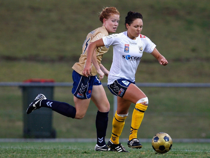 A soccer player wearing white, blue and yellow dribbles the ball past an opponent in blue and gold