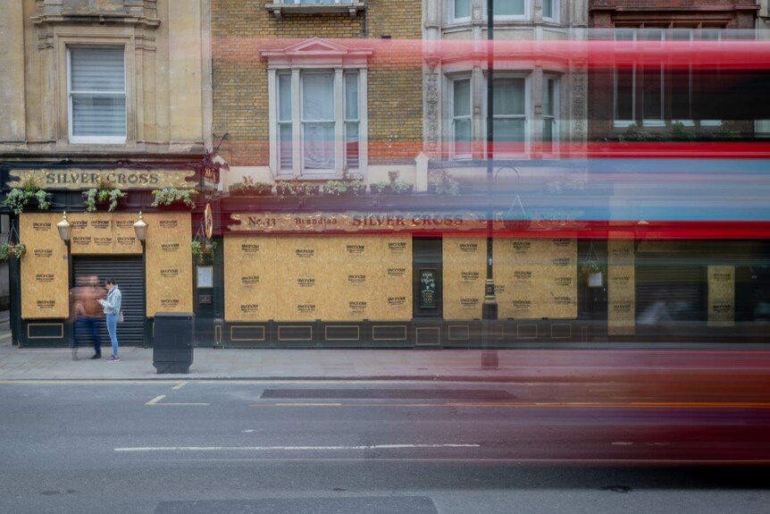 A time lapse photo of people walking past a boarded up pub while a double decker bus passes