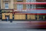 A time lapse photo of people walking past a boarded up pub while a double decker bus passes