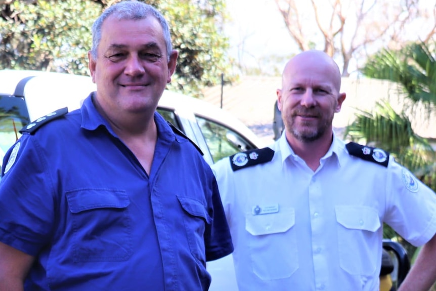 Two men smiling, one in blue and one in white uniform, stand next to a ute with a 'Marine Rescue' sign on it.
