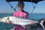 A woman in a pink top holds a large Spanish mackerel on a boat, smiling.