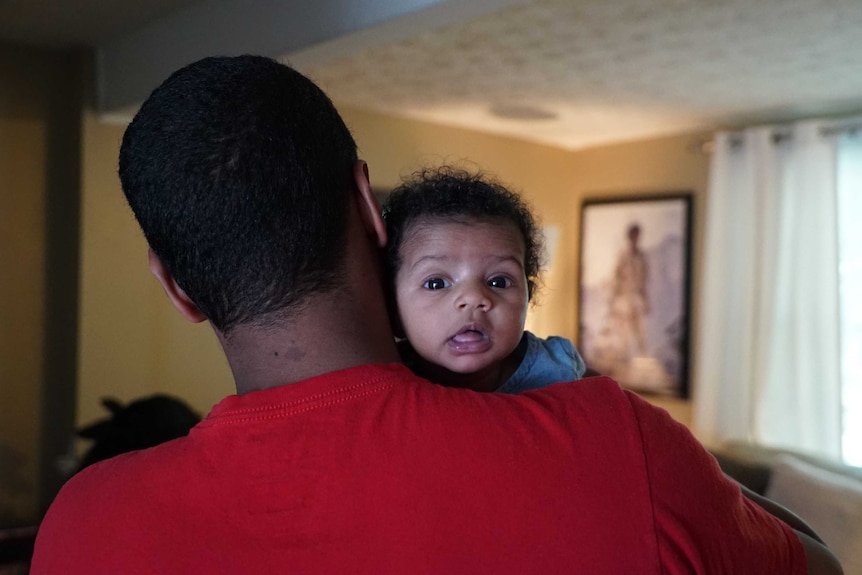 Baby Aroura stares over her father's shoulder