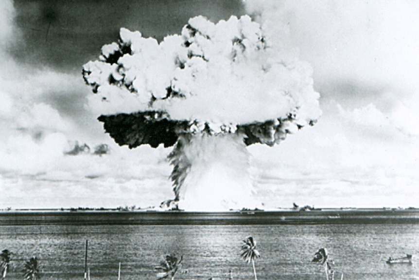 A black and white image of a mushroom cloud over water with palm trees in the foreground.