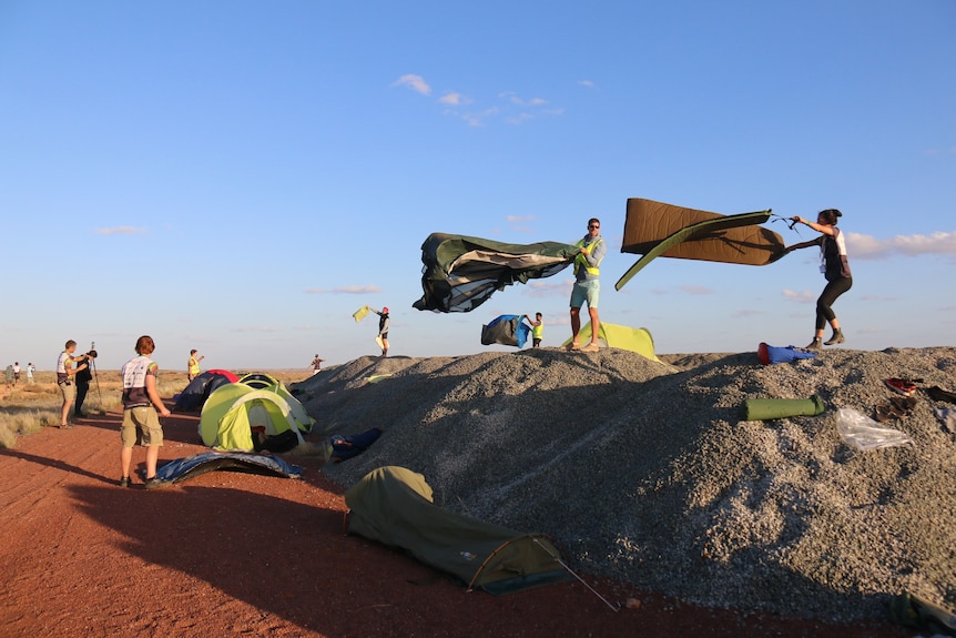People standing on gravel piles with sleeping mats and bags blowing in the wind.