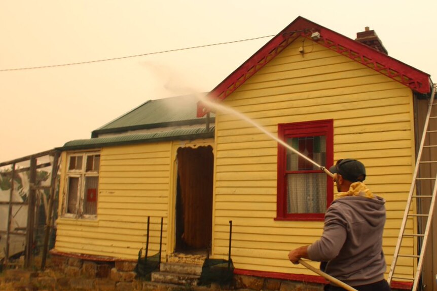 A man points a hose at a yellow weatherboard home, in the background there's smoke in the sky
