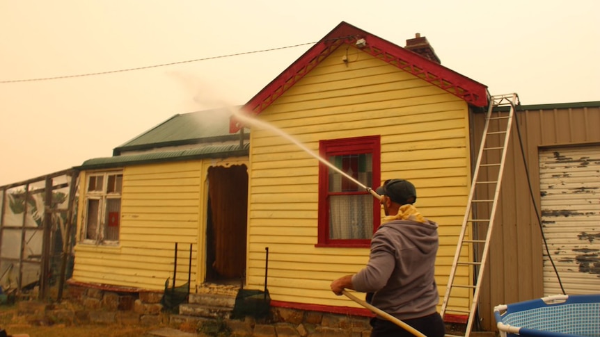 A man points a hose at a yellow weatherboard home, in the background there's smoke in the sky