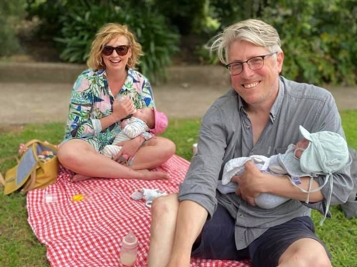 A man and a woman, each holding an infant, sit on a red and white checked picnic rug outside.