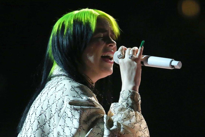 A woman with green hair sings in darkness on stage