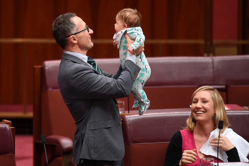 A man wearing a suit holds a baby girl as her mother looks on smiling.