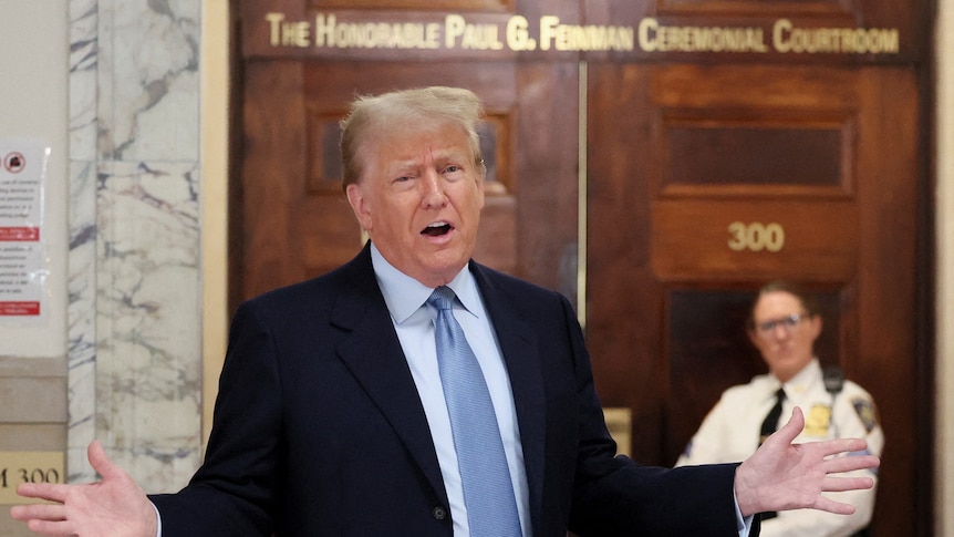 Donald Trump wearing a suit and blue tie standing with his arms spread apart with his mouth open