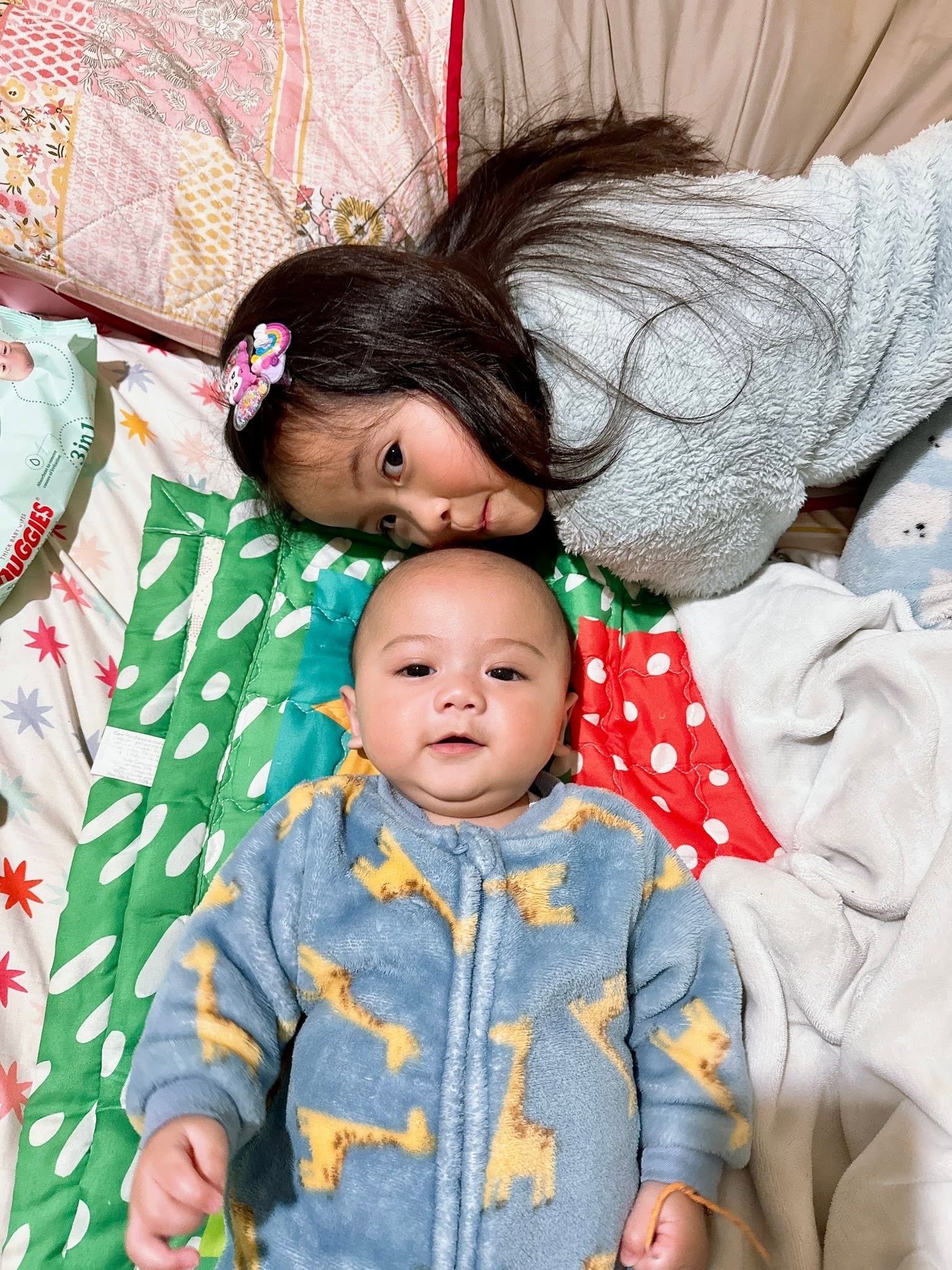 A young Asian girl with a grey sweater, smiling on a bed next to her baby brother smiling.