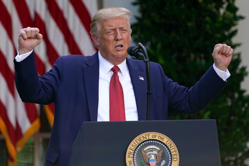 An elderly man in dark blue suit raises his arms in front of a microphone, with US flag behind him.