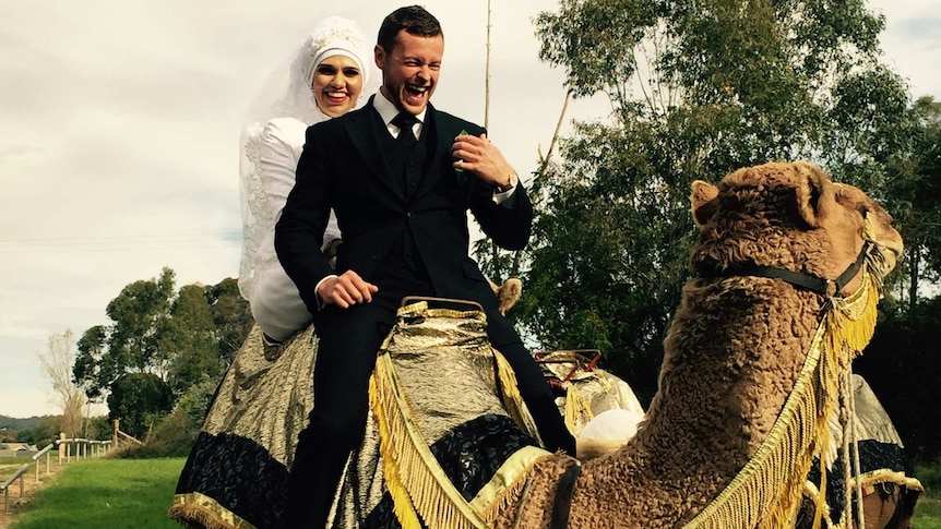 A woman in white wearing a veil sits behind a man on a camel. They are both laughing.