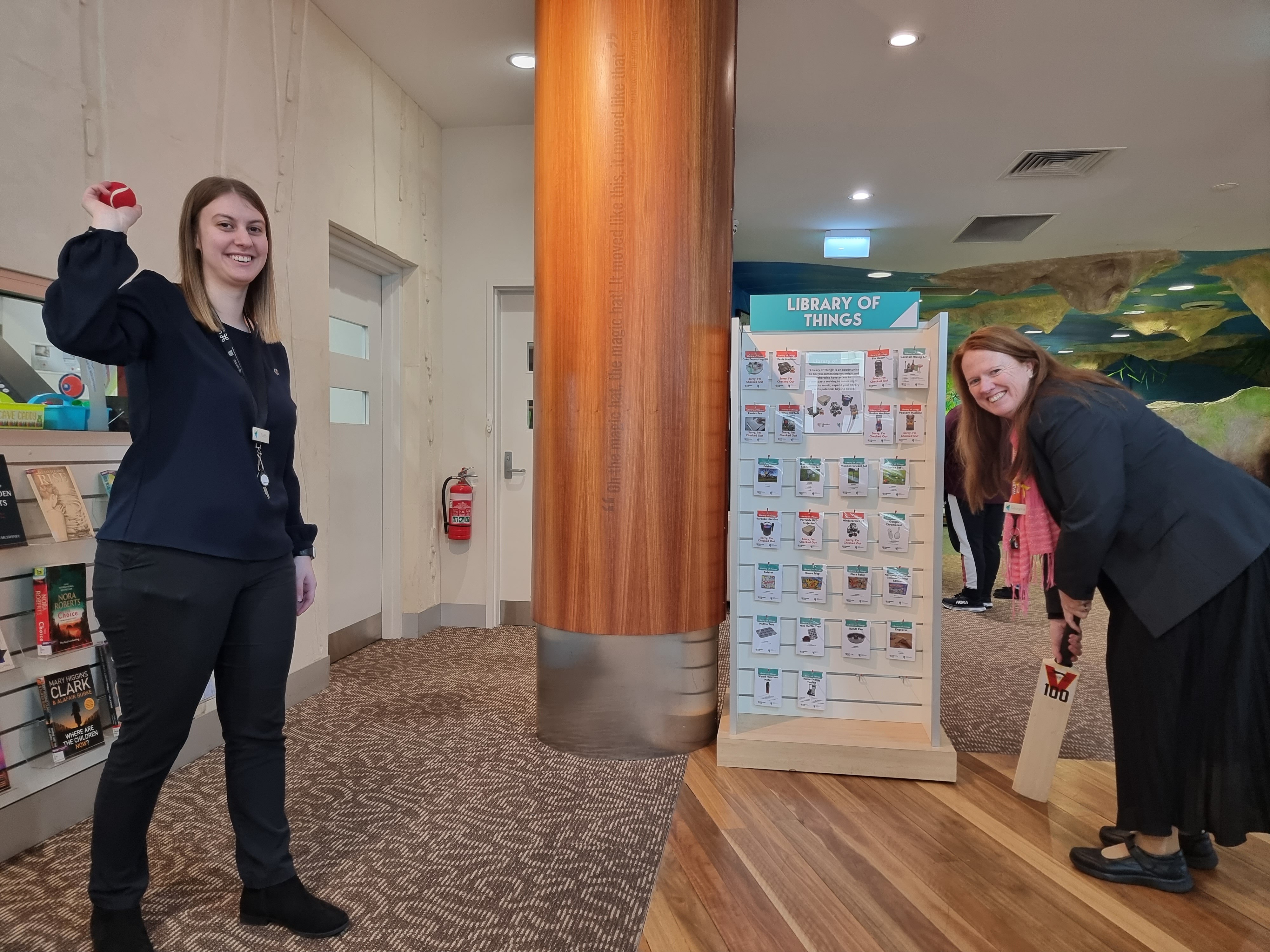 A woman holds up a red ball while another holds a cricket bat ready to play.