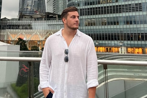 A young man with short brown hair and a short beard wears a white linen shirt and stands in front of glass buildings. 