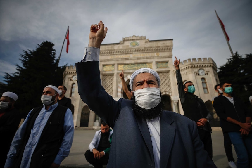 A group of men with face masks covering their mouth and beards stand facing the camera, with fingers in the air.