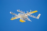 A white drone in the air with a package attached underneath it