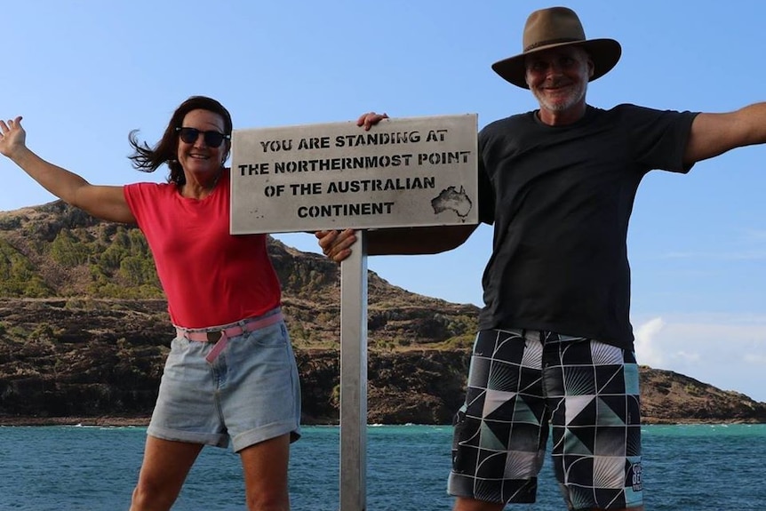Craig Dickman and a woman pose at the sign at the tip of Cape York with sea in the background.