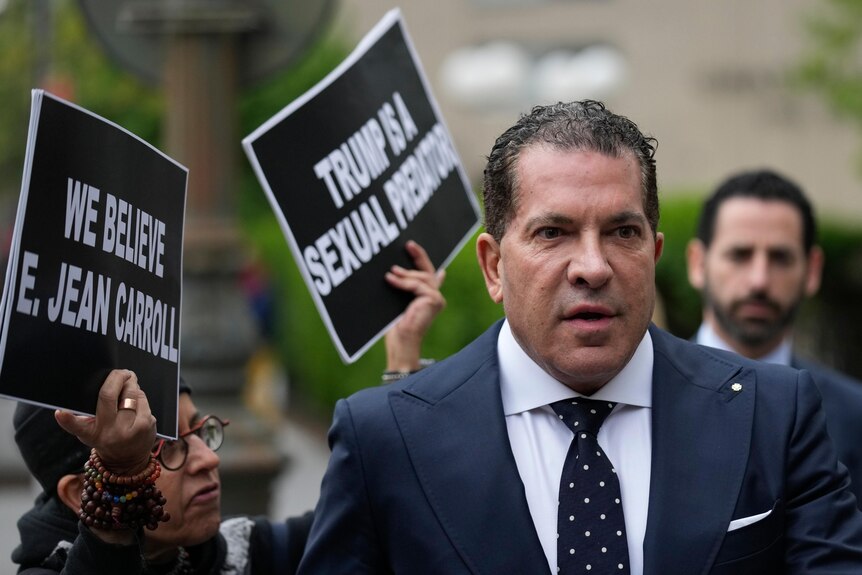 A man with short brown hair is pictured wearing a suit as he walks by a protester carrying signs in favour of E Jean Carroll.