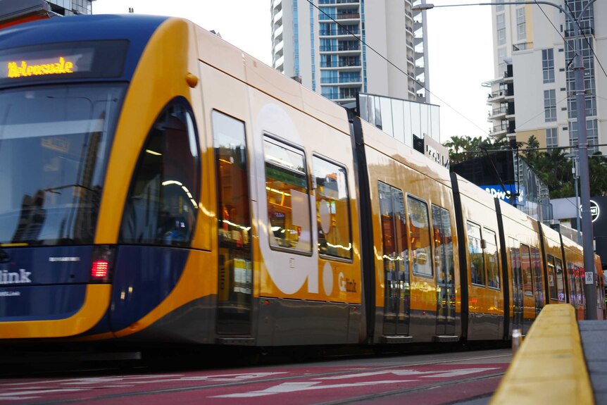 A yellow and blue light rail vehicle is stationary at a stop in Surfers Paradise. Tall buildings are in the distance