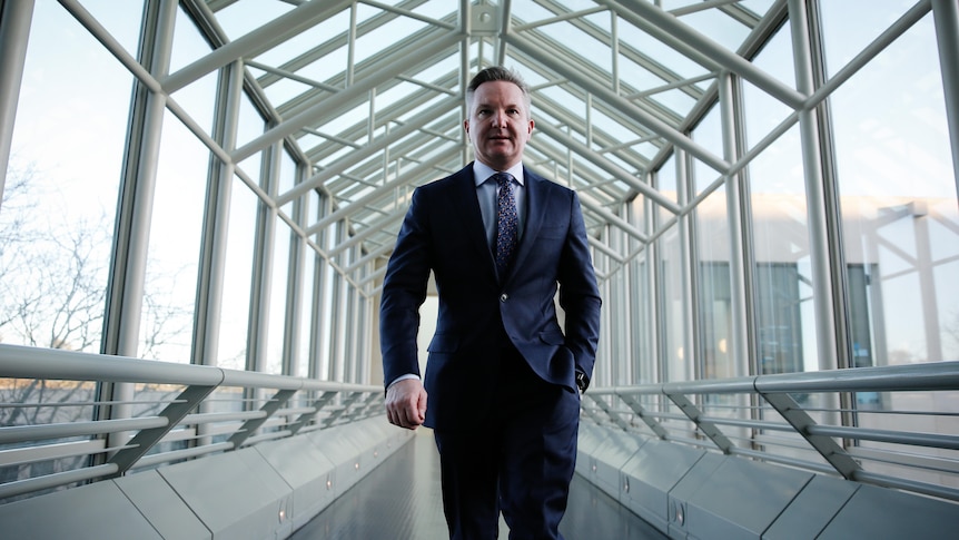 Bowen walks down a glass corridor with one hand in his pocket.