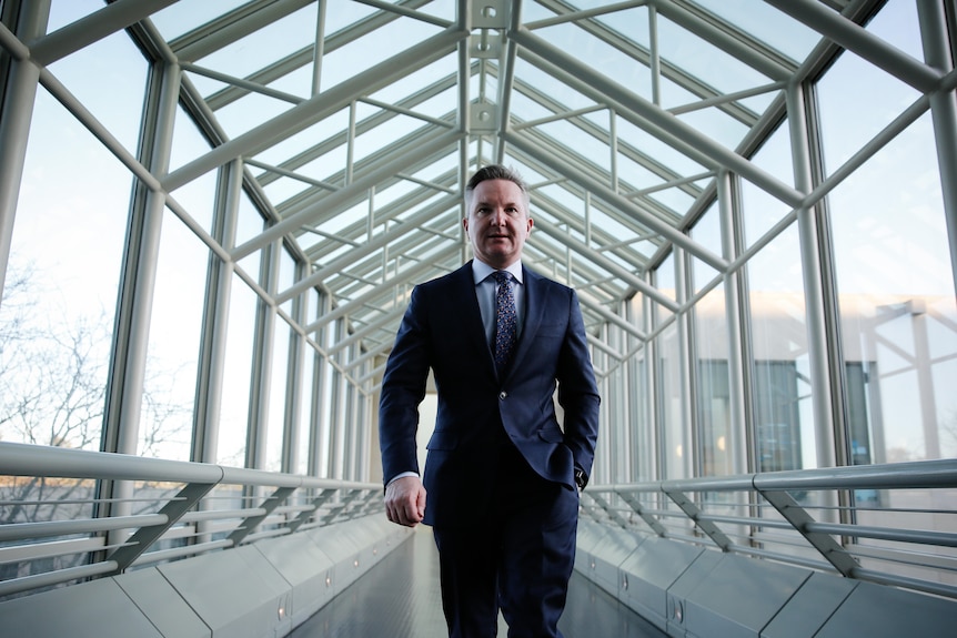 Bowen walks down a glass corridor with one hand in his pocket.