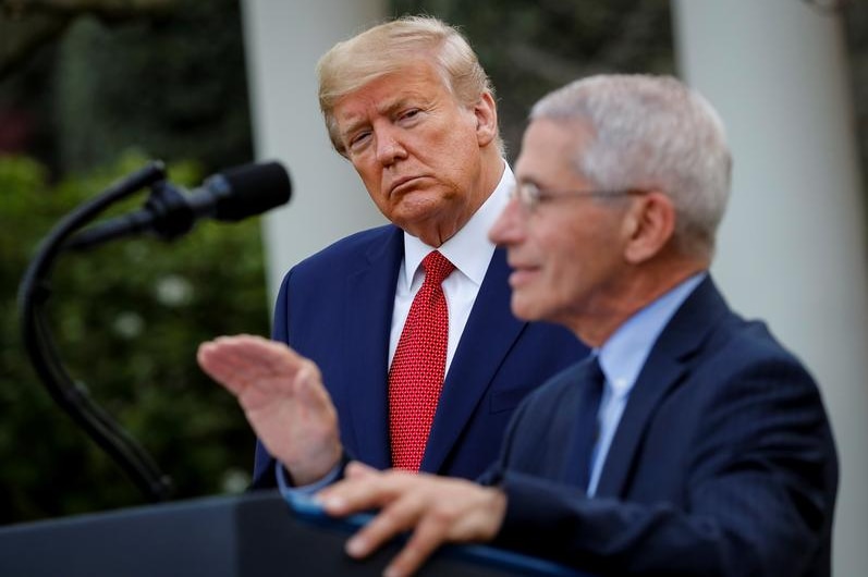 Anthony Fauci speaks at a podium while President Donald Trump watches.