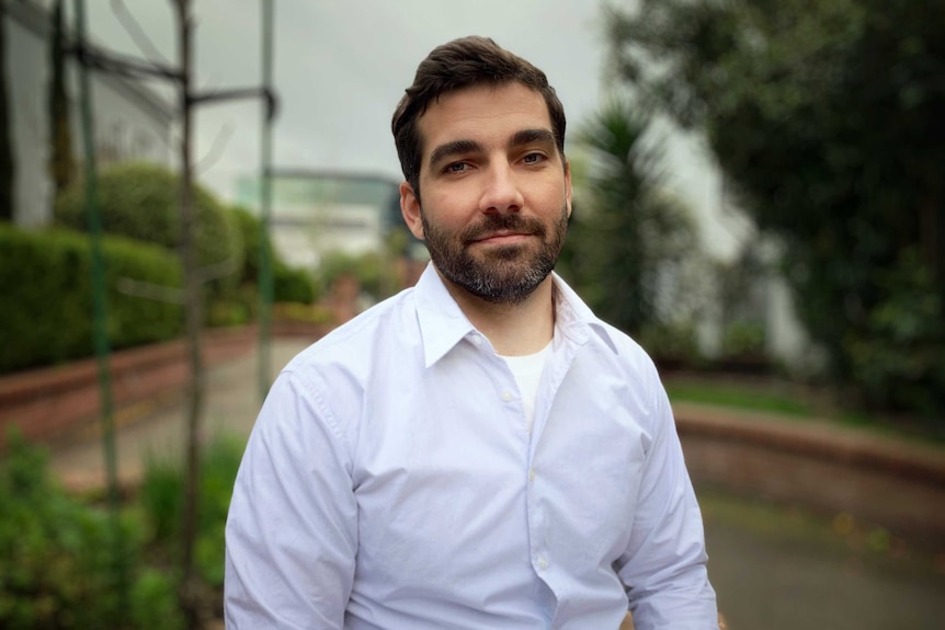 A man with dark hair and a short beard sits in a garden with a white collared shirt, looking into the camera.