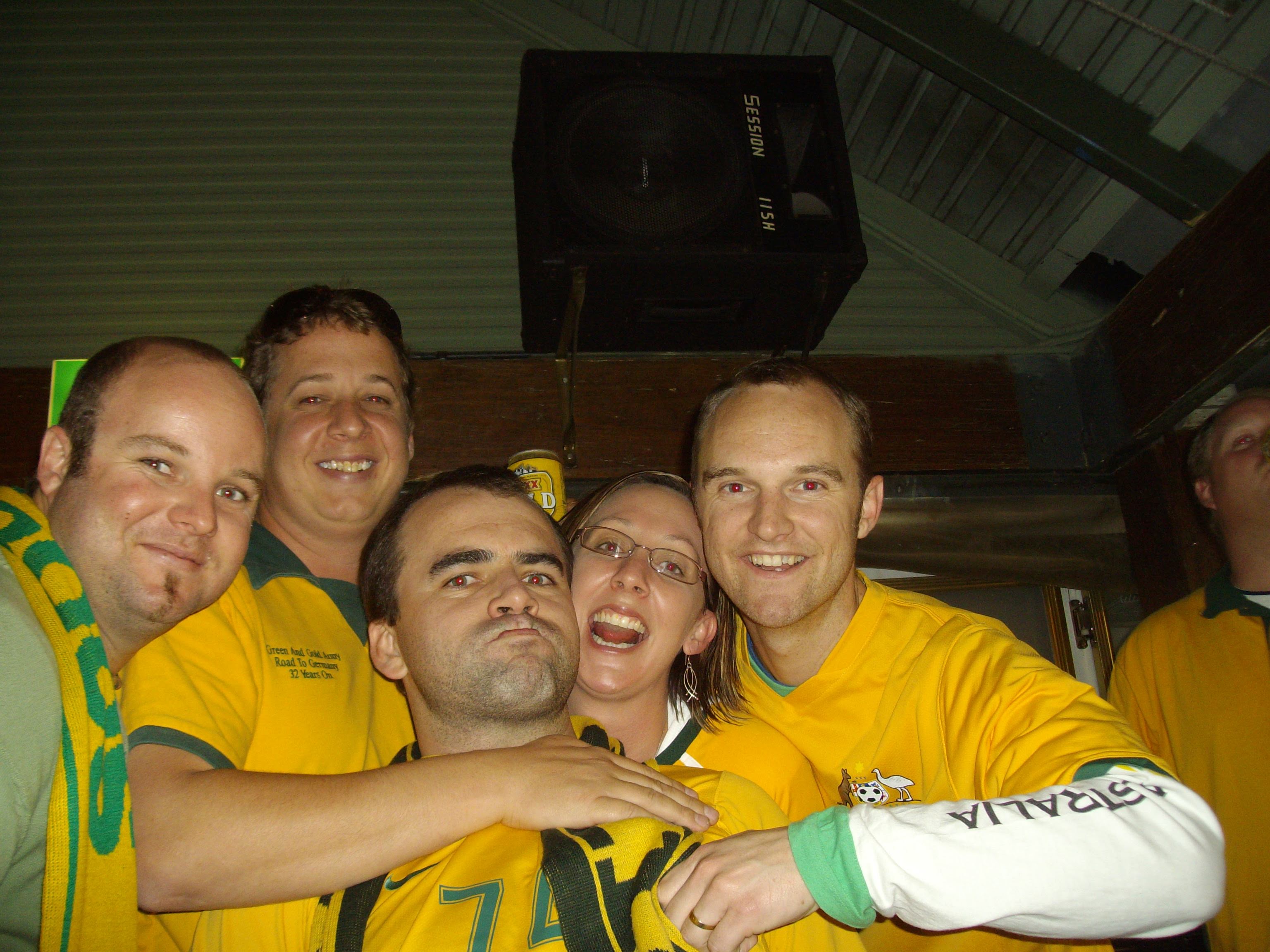 Four men and a woman, all smiling and wearing Socceroos jerseys as they cuddle up for a selfie.