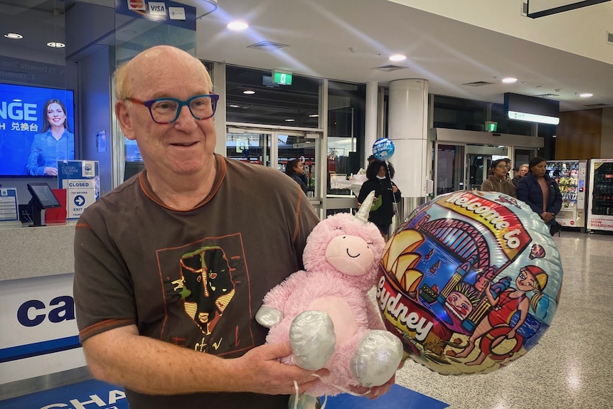 A man weaing glasses holds a pink stuffed unicorn and a colourful balloon as he waits in Sydney airport