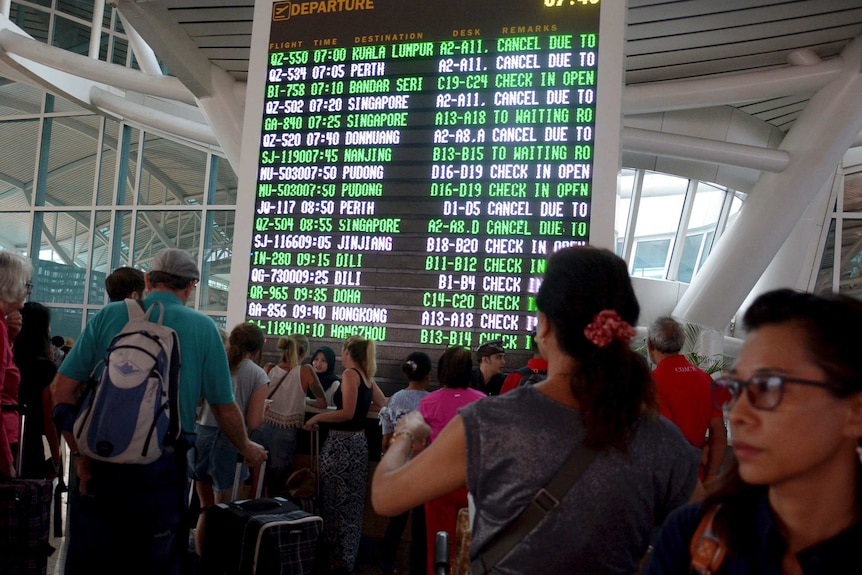 A flight information board shows cancelled flights at Bali's main airport, as passengers crowd around it with their luggage.