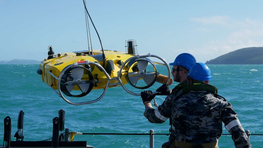 Navy sailors guide a yellow HMAS Huon into the ocean, which is lifted by wires