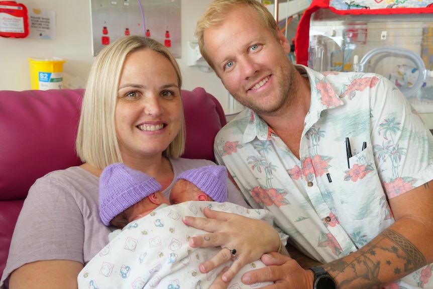 Justin and Tamara smiling at the camera with their hands on the twins, resting on Tamara's chest, purple beanies.