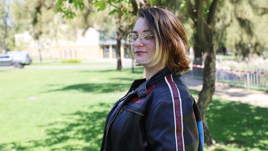 A brown haired girl wearing glasses and a black jacket with a camera looks at the camera in a park