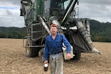 A cane harvesting contractor stands in front of his towering machine, with thermos in hand and esky over his shoulder