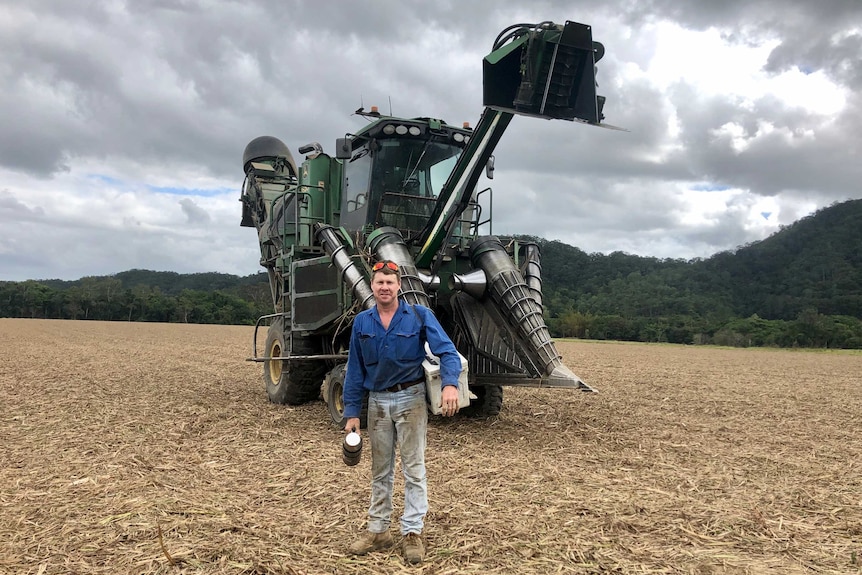 A cane harvesting contractor stands in front of his towering machine, with thermos in hand and esky over his shoulder