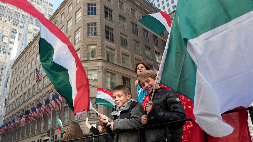 Two boys are waving large Italian flags while standing on a float. An office building with American flags is in the background.